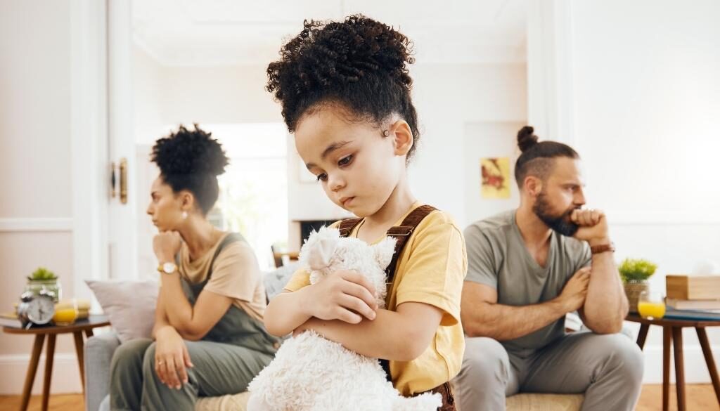 A couple looks frustrated while sitting opposite each other. Their child stands in the middle holding a stuffed animal with a sad look on her face.