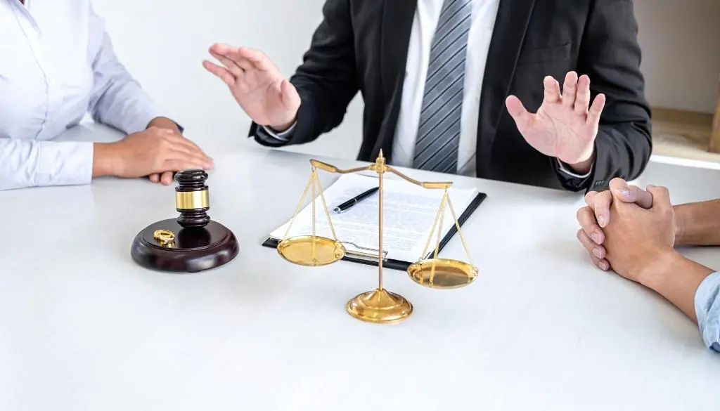 Man holds his hands up at a couple as a judge's gavel and scales of justice sit on the table in front of them.