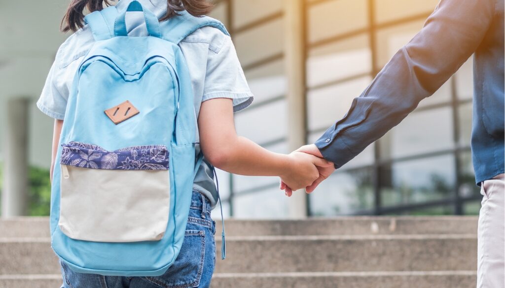 Child with a blue backpack holding an adult's hand in front of a school.