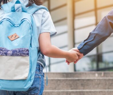 Child with a blue backpack holding an adult's hand in front of a school.