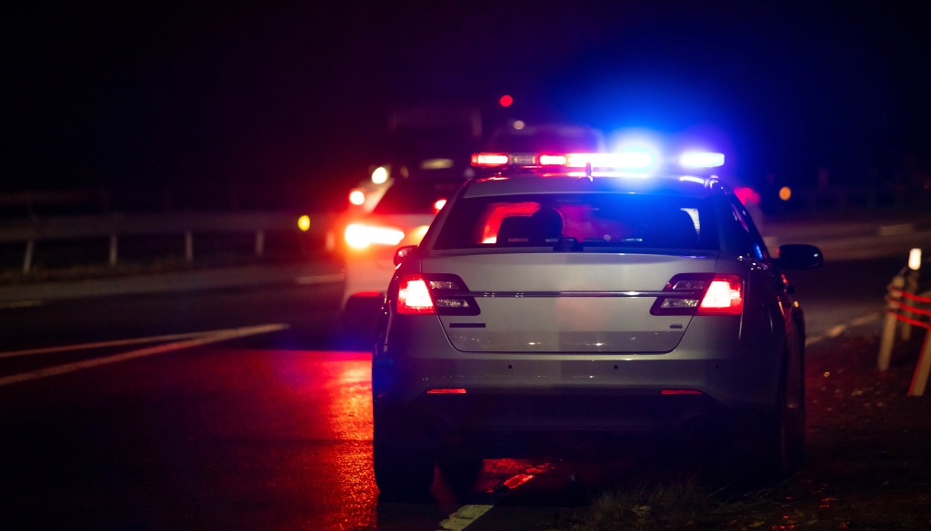 Police car with emergency lights on, pulled over at night on a road, with another vehicle in the background, suggesting a traffic stop or law enforcement action.