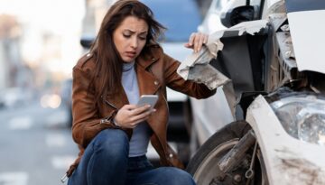 Woman examining damage to her car after an accident, holding her phone and looking concerned.