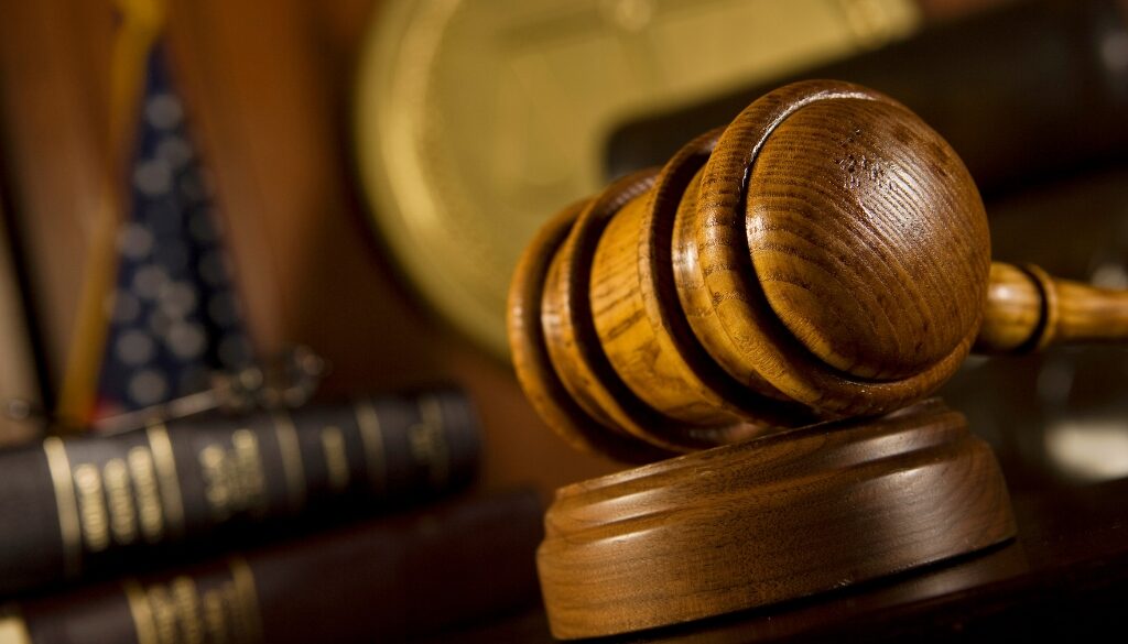 A wooden judge's gavel resting on its block in a courtroom, with legal books, an American flag, and scales of justice blurred in the background.