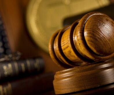 A wooden judge's gavel resting on its block in a courtroom, with legal books, an American flag, and scales of justice blurred in the background.