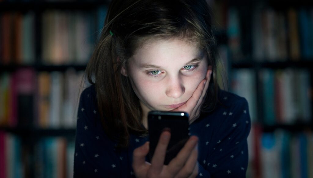 A young girl looking at a smartphone with a concerned expression, sitting in a dimly lit room with bookshelves in the background.