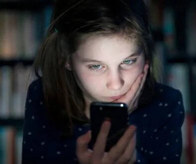 A young girl looking at a smartphone with a concerned expression, sitting in a dimly lit room with bookshelves in the background.