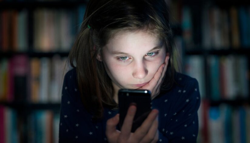 A young girl looking at a smartphone with a concerned expression, sitting in a dimly lit room with bookshelves in the background.