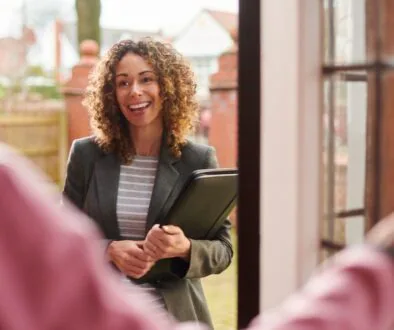 A professional landlord meeting a tenant at the door of a residential property, holding a folder and smiling warmly.