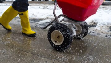 Person wearing bright yellow boots spreads salt on an icy sidewalk using a red spreader.