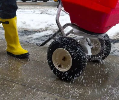 Person wearing bright yellow boots spreads salt on an icy sidewalk using a red spreader.