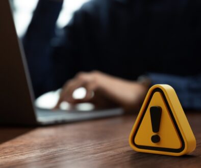 A yellow warning sign with an exclamation mark sits on a wooden desk in front of a person working on a laptop.