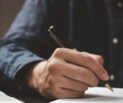 A close-up of a person's hand holding a black and gold pen while filling out a form on a desk. The individual is wearing a dark-colored, button-up shirt.