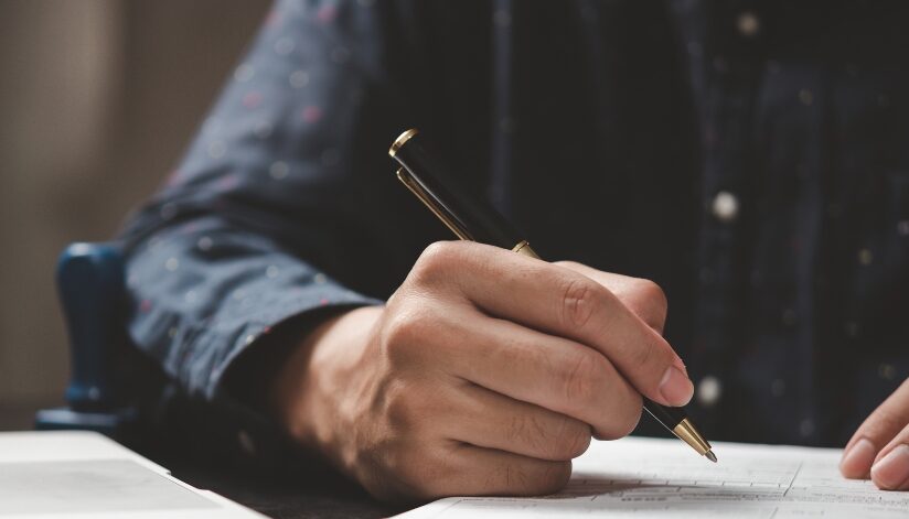 A close-up of a person's hand holding a black and gold pen while filling out a form on a desk. The individual is wearing a dark-colored, button-up shirt.