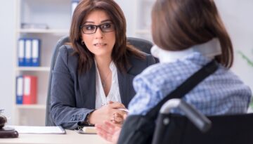 A woman in a wheelchair wearing a neck brace speaks with a personal injury lawyer during a legal consultation in an office setting.