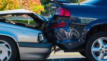 A silver car rear-ended a black sedan on a sunny street, showing visible damage to both vehicles.