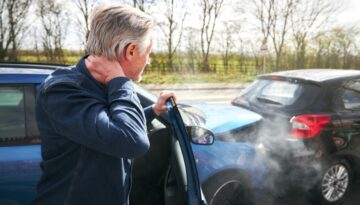 Man holding his neck in pain after a rear-end car accident, standing next to his vehicle with visible collision damage and smoke.