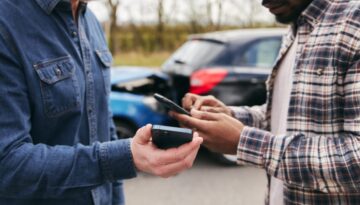 Two men exchange information on smartphones after a rear-end car accident involving out-of-state visitors.