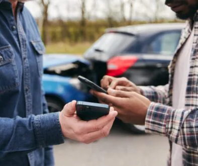 Two men exchange information on smartphones after a rear-end car accident involving out-of-state visitors.