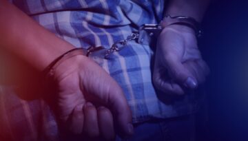 A person with handcuffs behind their back and red and blue police lights in the distance.