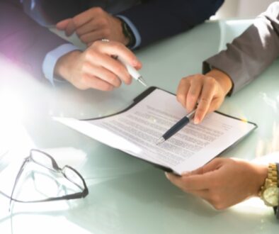 Two people sit across a glass table reviewing a legal document, each holding a pen and pointing to sections of the paperwork, with a pair of eyeglasses resting nearby.