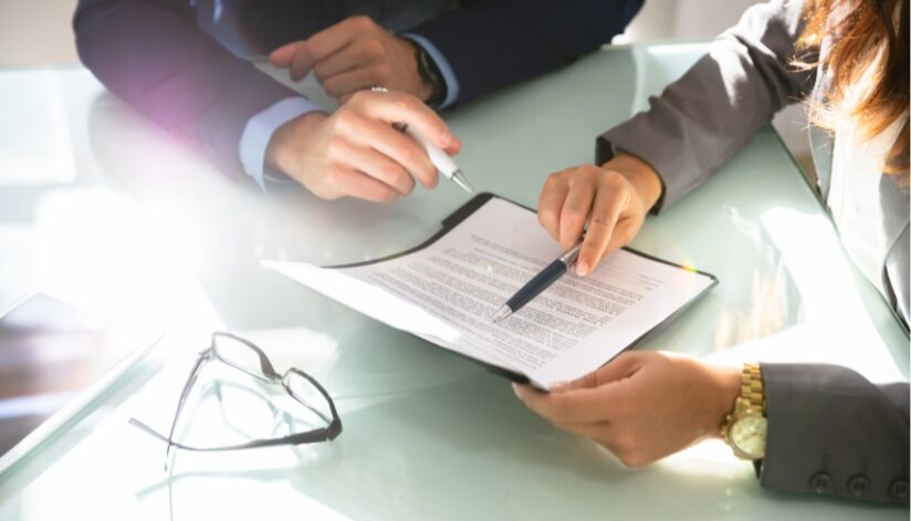 Two people sit across a glass table reviewing a legal document, each holding a pen and pointing to sections of the paperwork, with a pair of eyeglasses resting nearby.