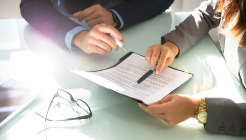 Two people sit across a glass table reviewing a legal document, each holding a pen and pointing to sections of the paperwork, with a pair of eyeglasses resting nearby.