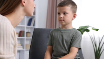 A young boy sits on a chair while an adult woman leans toward him, speaking calmly in a bright, modern living room setting.