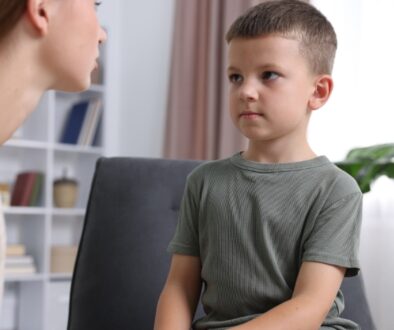 A young boy sits on a chair while an adult woman leans toward him, speaking calmly in a bright, modern living room setting.