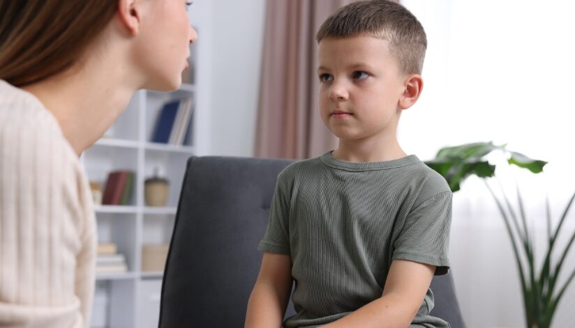 A young boy sits on a chair while an adult woman leans toward him, speaking calmly in a bright, modern living room setting.
