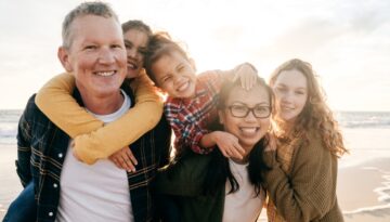 A smiling blended family with two parents and three children standing together on a beach at sunset.