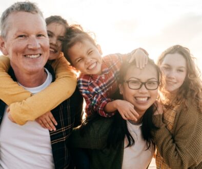 A smiling blended family with two parents and three children standing together on a beach at sunset.