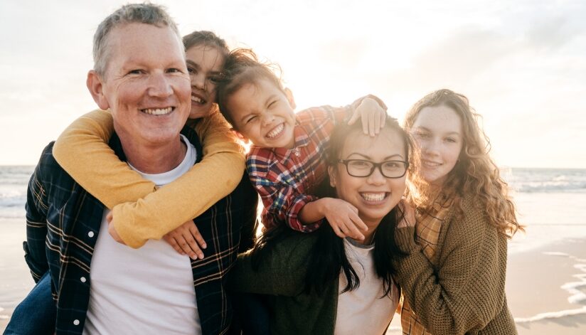 A smiling blended family with two parents and three children standing together on a beach at sunset.