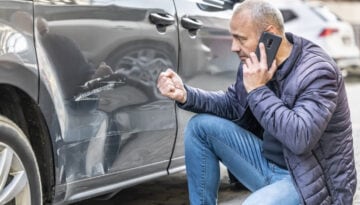 Man kneeling and assessing damages to his car while talking on a cell phone.