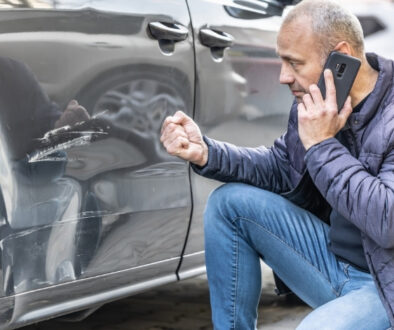 Man kneeling and assessing damages to his car while talking on a cell phone.