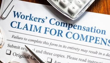 Close-up of a workers’ compensation claim form with a pen, calculator, paper clip, and eyeglasses on a wooden desk.
