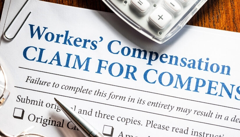Close-up of a workers’ compensation claim form with a pen, calculator, paper clip, and eyeglasses on a wooden desk.