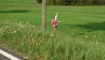 Roadside memorial cross with flowers beside a rural road and grassy field.