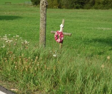Roadside memorial cross with flowers beside a rural road and grassy field.