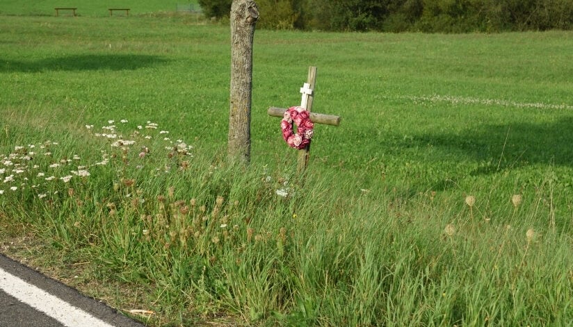 Roadside memorial cross with flowers beside a rural road and grassy field.