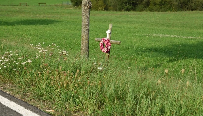Roadside memorial cross with flowers beside a rural road and grassy field.